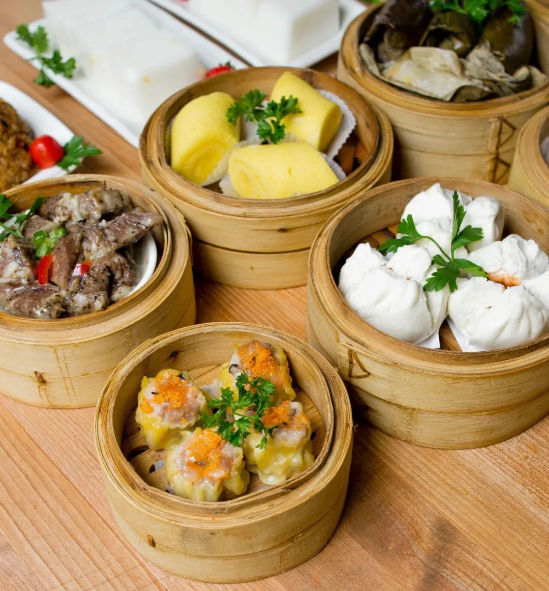 木桌上摆竹制蒸笼，内有虾饺、烧卖、叉烧包等中式点心，点缀绿叶，展现传统美食色彩与质感，氛围温馨。