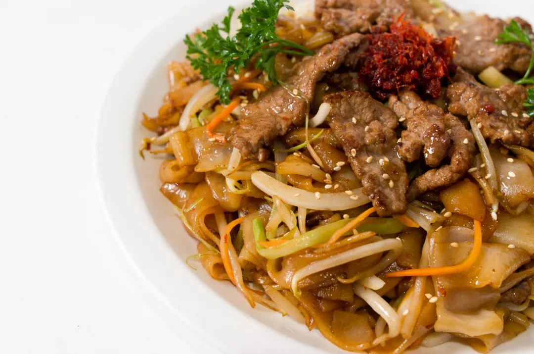 白盘盛香煎牛肉河粉，嫩牛肉盖河粉，夹豆芽、胡萝卜丝、西芹条，撒白芝麻与欧芹，顶部有红酱，色泽鲜亮诱人。