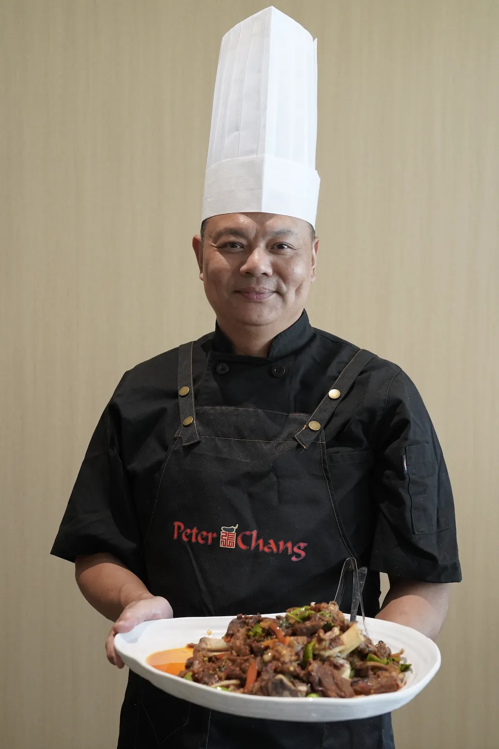 戴白高帽、穿“Peter Chang”红字黑服的厨师双手捧菜，盘中有肉蔬，背景浅色墙面，突出厨师与美食的专业感。