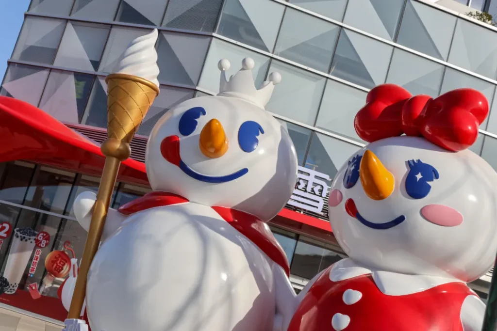 画面展示蜜雪冰城门店外的两尊卡通雪人雕塑，左侧雪人戴皇冠、持金色冰淇淋权杖，右侧雪人配红色大蝴蝶结，背景是品牌标志性玻璃建筑，色彩鲜亮、形象活泼，具品牌辨识度。