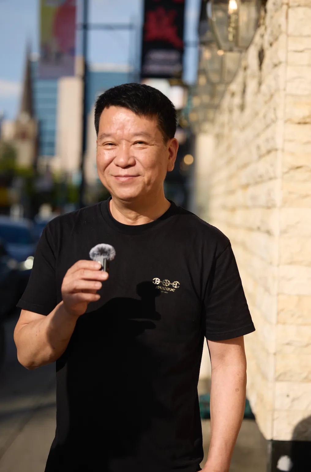 黑衣男子面带微笑，手持物品站石墙建筑前，背景是城市街道，阳光洒落，氛围轻松自然，展现随性状态。