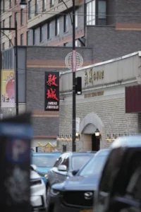 唐人街街道近处停汽车，远处左侧有“敦城 ASIAN JEWELS”黑招牌，右侧建筑金色文字，展现城市唐人街街景风貌。
