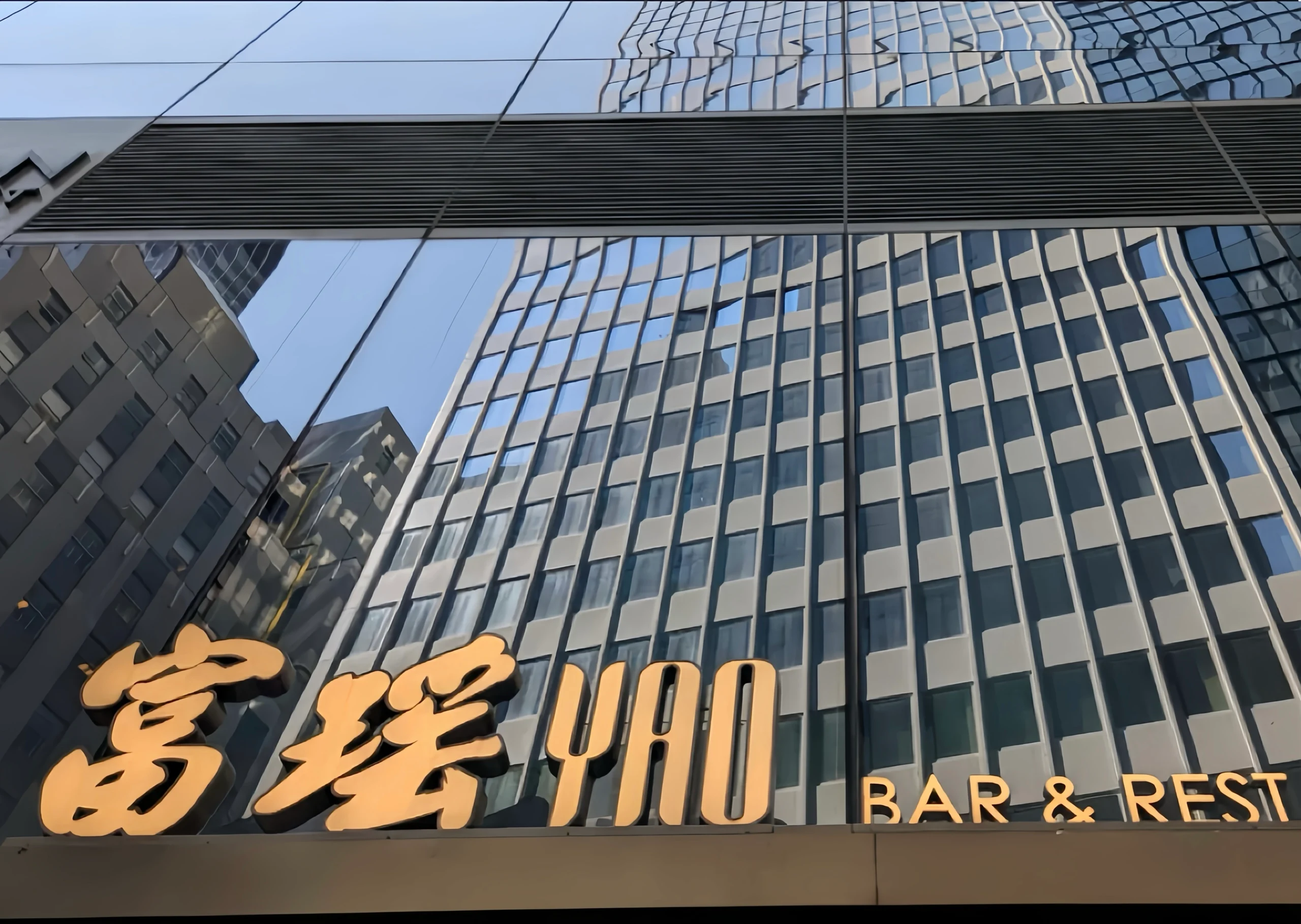 富瑶华尔街旗舰店"YAO BAR & RESTAURANT"金色立体字门头，安装于万豪酒店大理石外立面。背景玻璃幕墙反射金融区摩天楼群，下方"BAR"字样点明酒吧功能，印证"高端粤菜进驻金融核心区"战略。