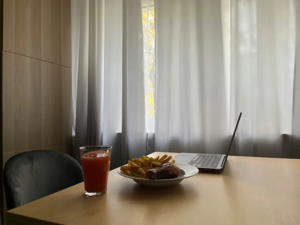 浅木桌上摆红色饮品、薯条肉盘与打开的笔记本，背景白窗帘透出室外黄叶，整体氛围轻松闲适，展现边吃边办公的日常场景。