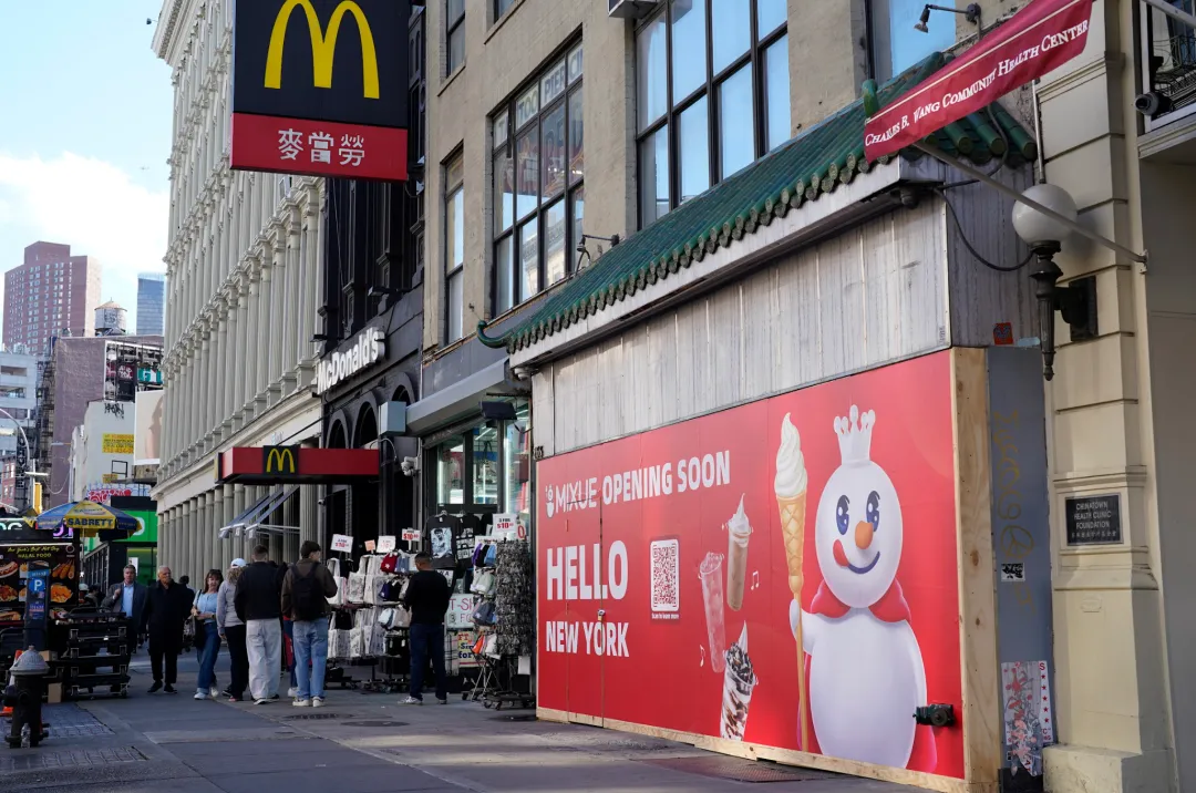纽约街景，右侧建筑外墙写“蜜雪冰城即将开业”，左侧有麦当劳标识，行人往来，呈现多元商业氛围。