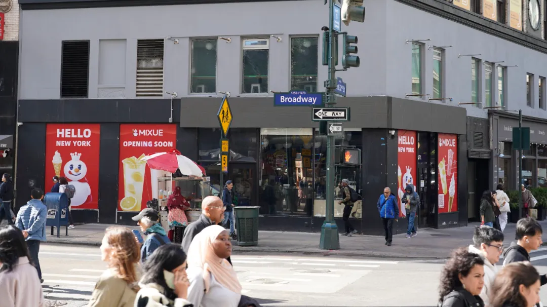 纽约百老汇街角蜜雪冰城门店，外墙红广告写“HELLO NEW YORK”“MIXUE OPENING SOON”，行人穿梭，旁有交通信号灯。