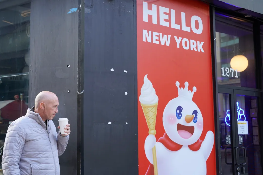 左侧穿浅灰羽绒服拿杯子的光头男，右侧蜜雪冰城红招牌写“HELLO NEW YORK”，有雪人形象，门牌号1271，为纽约店外景。